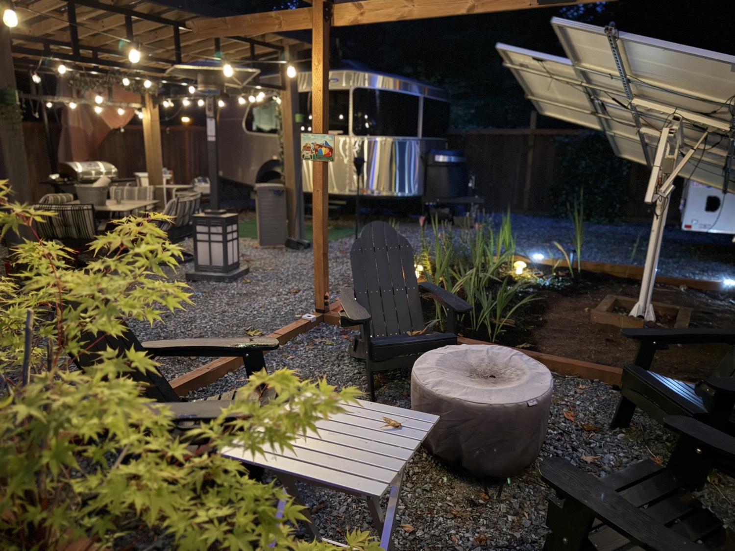 Black Adirondack chairs on gravel, a Japanese maple, and a silver trailer under string lights